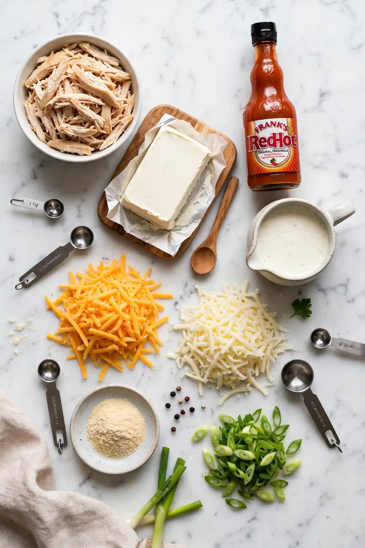 All ingredients for buffalo chicken dip laid out on white marble — shredded chicken, cream cheese, hot sauce, ranch, cheddar, and green onions — buffalo chicken dip ingredients