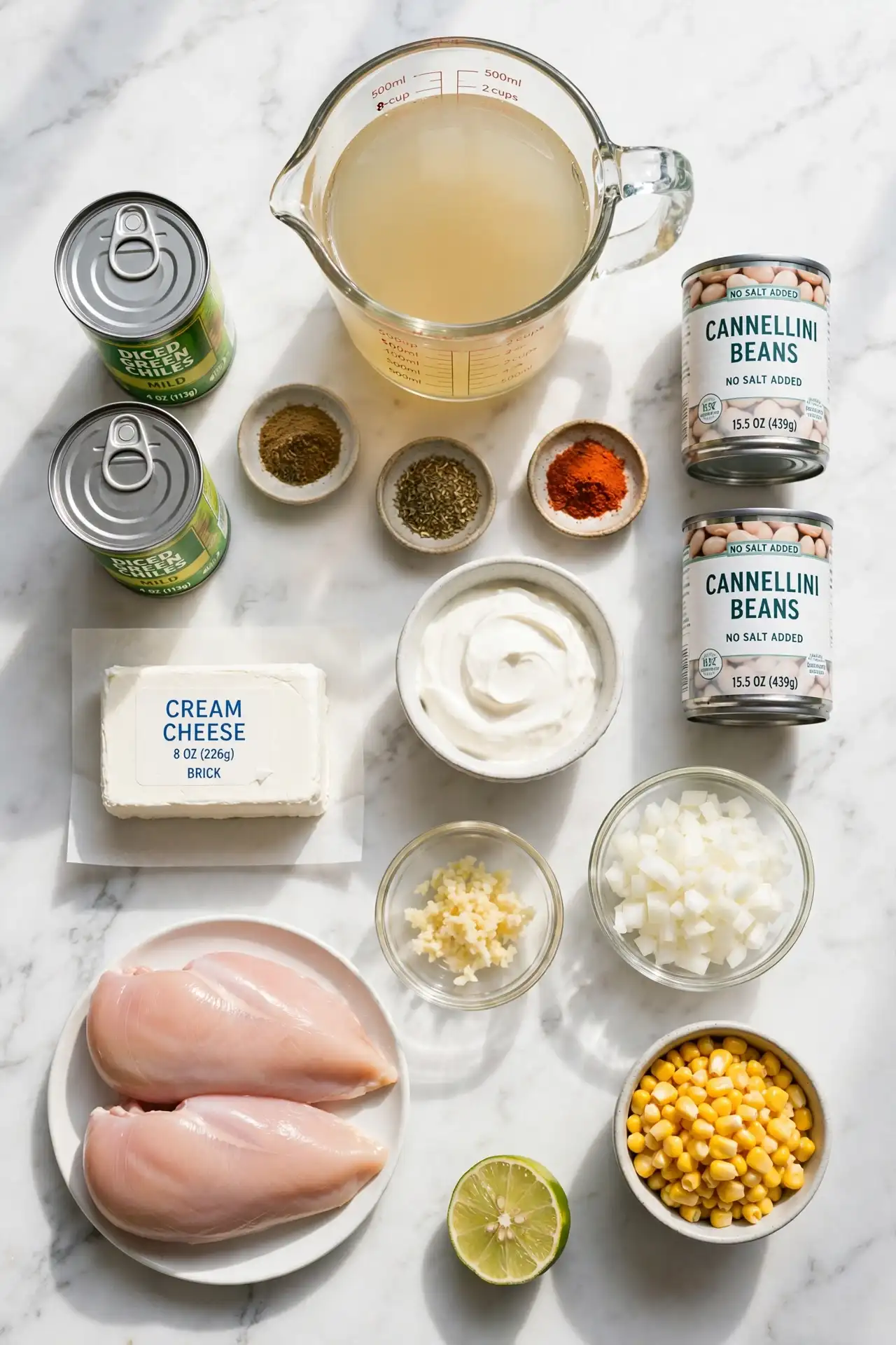 All ingredients for white chicken chili laid out on white marble, including chicken breasts, canned beans, green chiles, cream cheese, and spices — white chicken chili ingredients