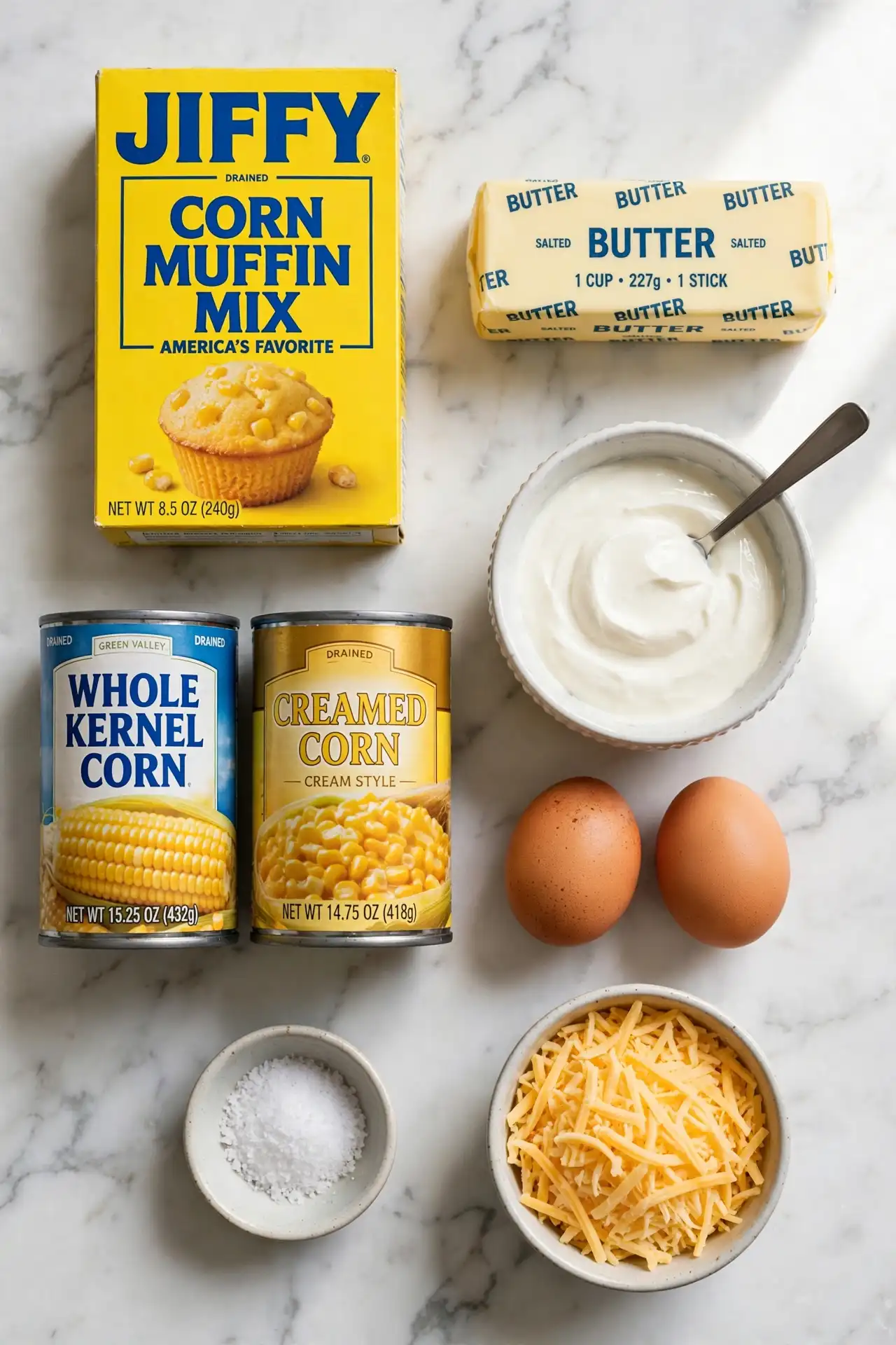 All ingredients for corn casserole laid out flat on white marble — corn casserole ingredients including creamed corn, Jiffy mix, sour cream, and butter